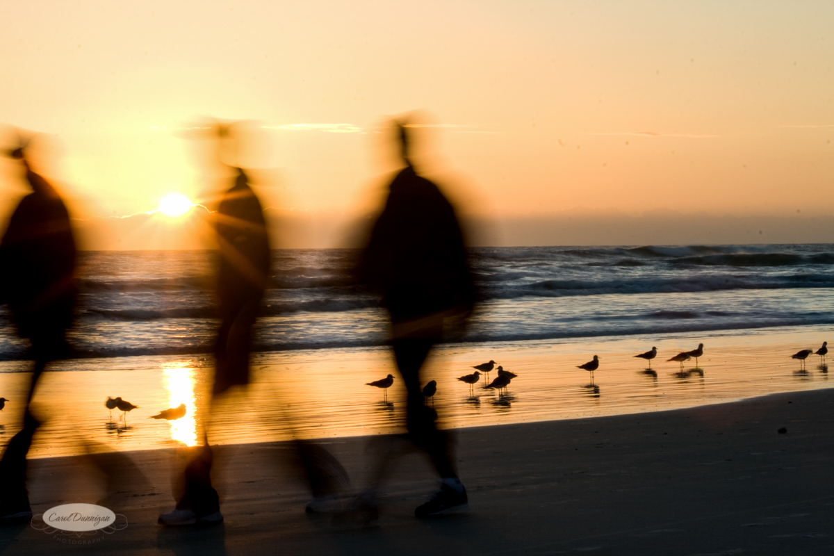 A slow motion view of two people walking on Daytona Beach in Florida. It is sunrise, and you can see birds and the ocean in the background.