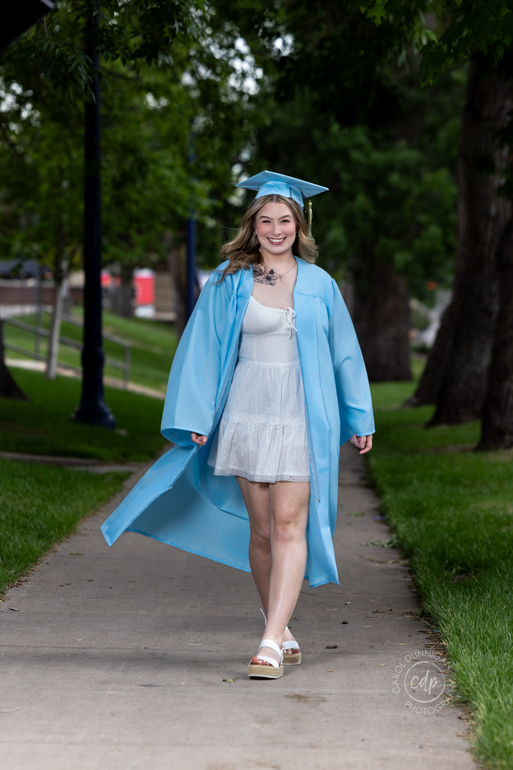 A Greeley West high school graduate is walking down a sidewalk with her cap and gown on during her cap and gown session with carol dunnigan photography in greeley colorado