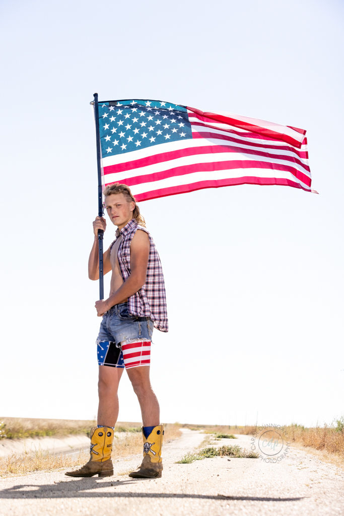 A high school senior guy is posing for his senior pictures with Carol Dunnigan Photography in Berthoud, Colorado. He's holding an American flag, wearing a cut off jean jacket, jean shorts with the American flag, and cowboy boots. He also has a mullet hair cut.