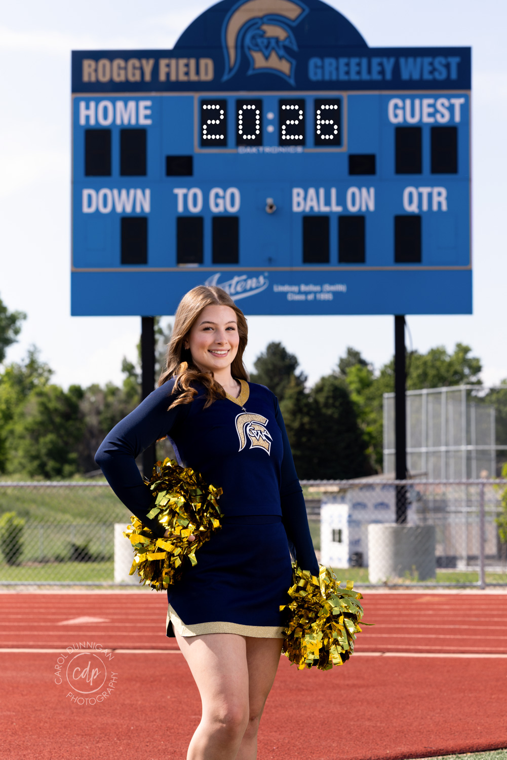 greeley high school cheerleader during her senior pictures with the roggy field greeley west score sign behind her and the numbers 2026 photographed by carol dunnigan photography