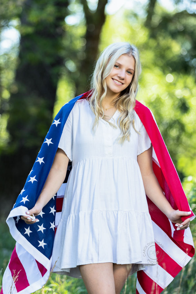 High School senior posing in a tree filled area wearing a white dress and an american flag over her shoulders. Taken in Fort Collins Colorado by Carol Dunnigan Photography.