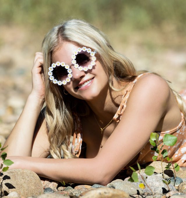 High school senior picture of a girl lying on river rock with daisy sunglasses on being photographed in Fort Collins by Carol Dunnigan Photography.