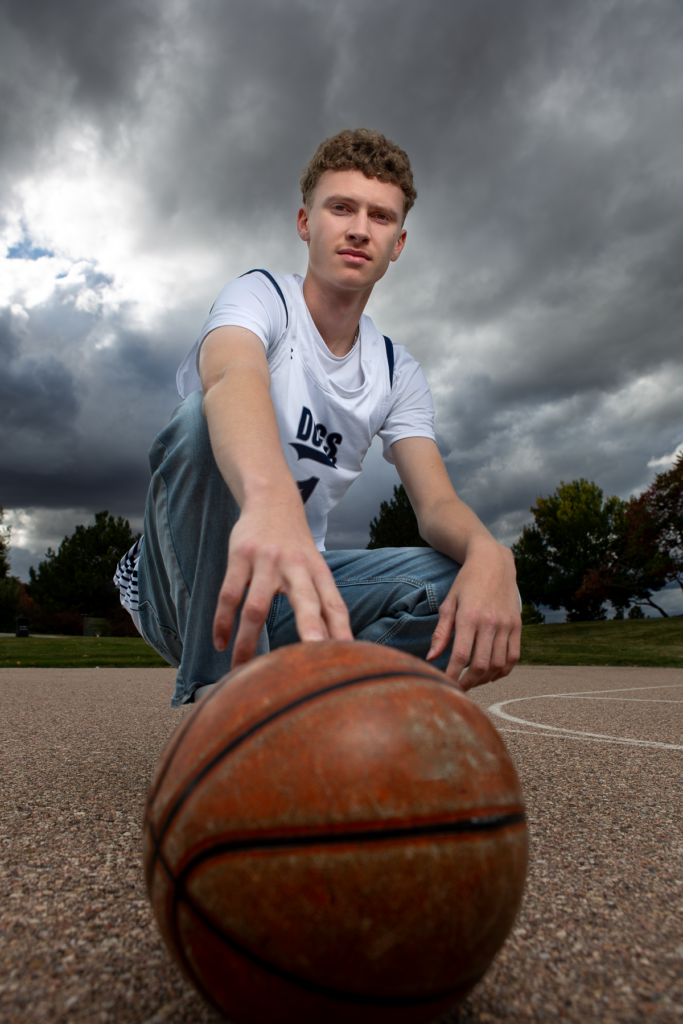 High school senior basketball pictures in Greeley, Colorado. University high school boy is squatting with his finger tips on a basketball with a dark sky above him.