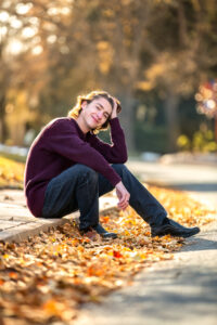Senior boy is posing in the Fall in Greeley Colorado. He's sitting on a curb with yellow leaves on the ground.