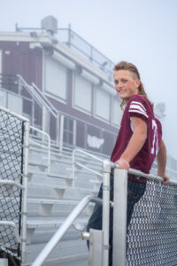 Berthough high school football player in Colorado is standing with his back against the bleacher railing and looking to the side and smiling to the photographer who is taking his senior pictures. Carol Dunnigan Photography.