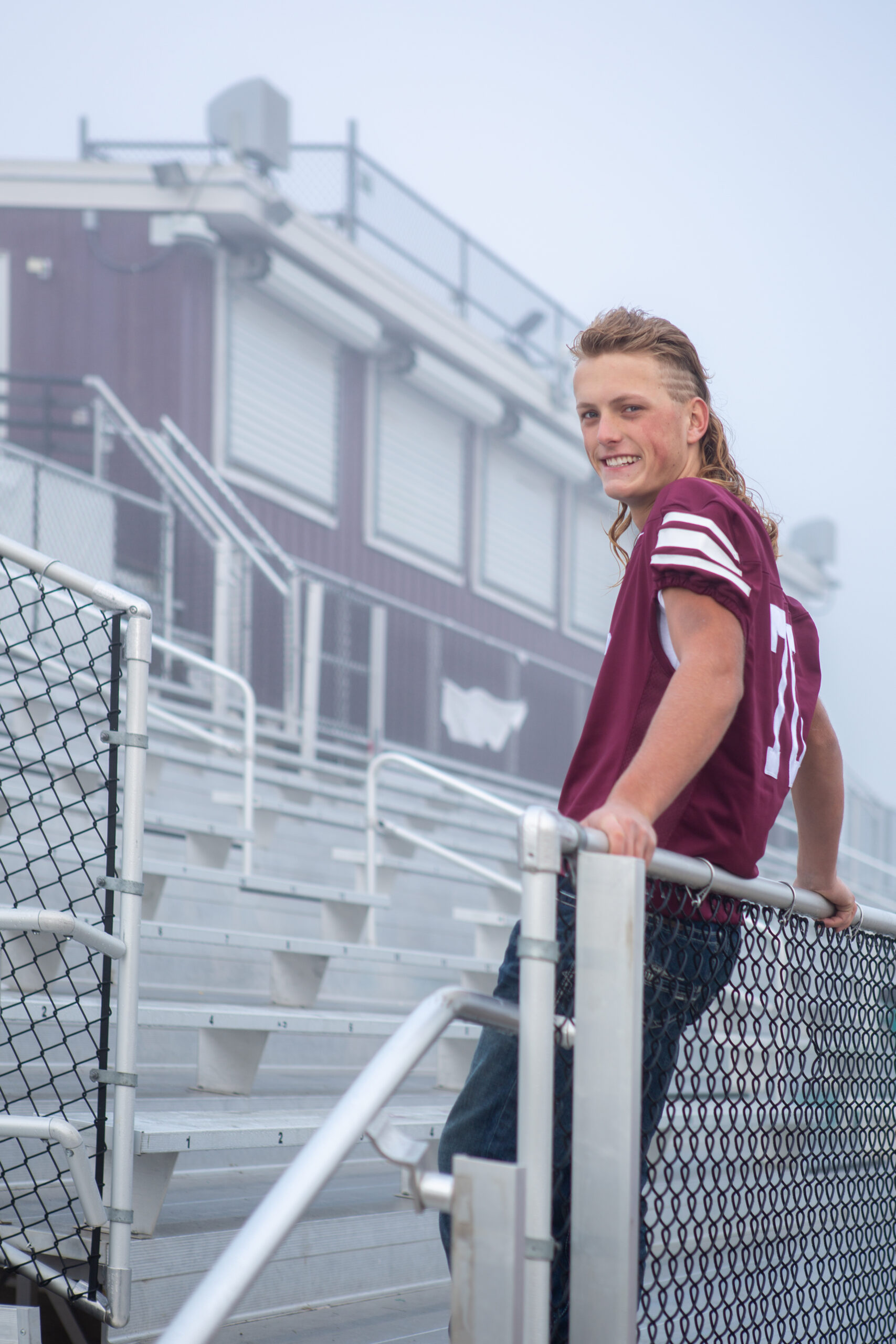 A Berthoud Colorado high school football player is casually in the bleachers, leaning against the railing with his back and smiling at the photographer during his senior session with Carol Dunnigan Photography.