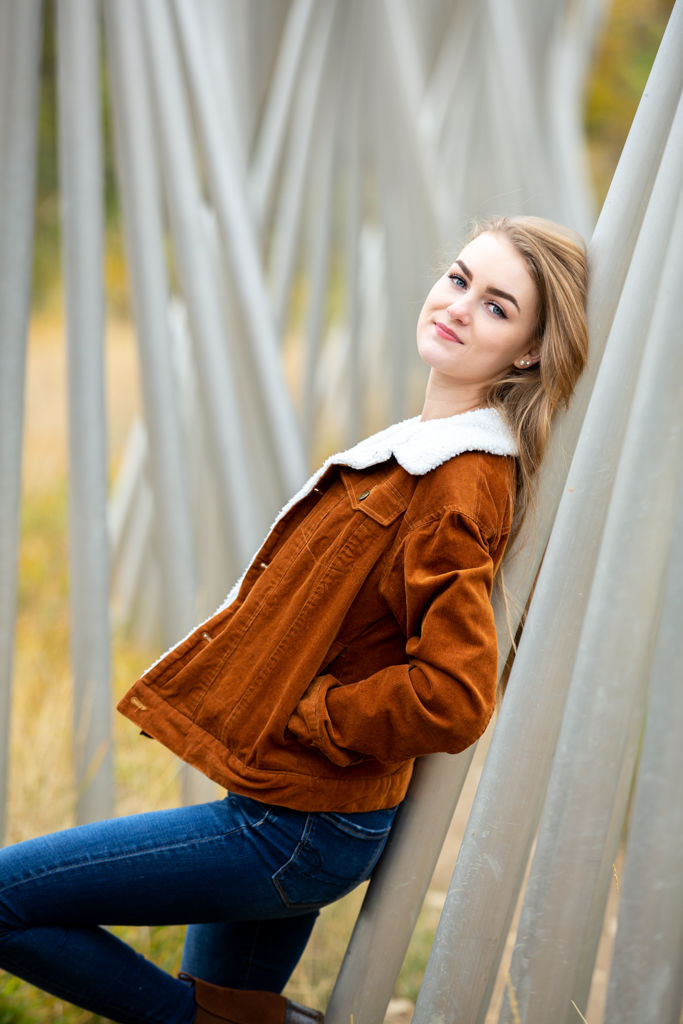 High school senior wearing a brown jacket layered over a casual outfit for fall senior pictures in Evans Colorado.