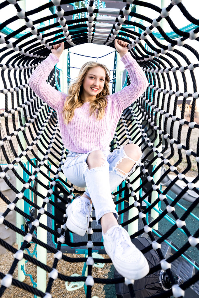 High school senior in Greeley Colorado wearing a pink sweater, ripped jeans, and white shoes on a playground climbing net.