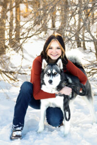 Senior girl in Evans Colorado is posing in the snow with her Siberian Husky. She's squatting down next to him, hugging him and smiling.