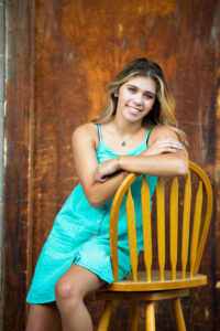 A high school senior girl is posing on a wood chair, against a good door in Fort Collins, Colorado during her senior session with Carol Dunnigan Photography.
