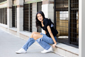 High school senior girl posing for senior pictures outside of a parking garage, sitting on a ledge and leaning forward in Fort Collins Colorado by Carol Dunnigan Photography.