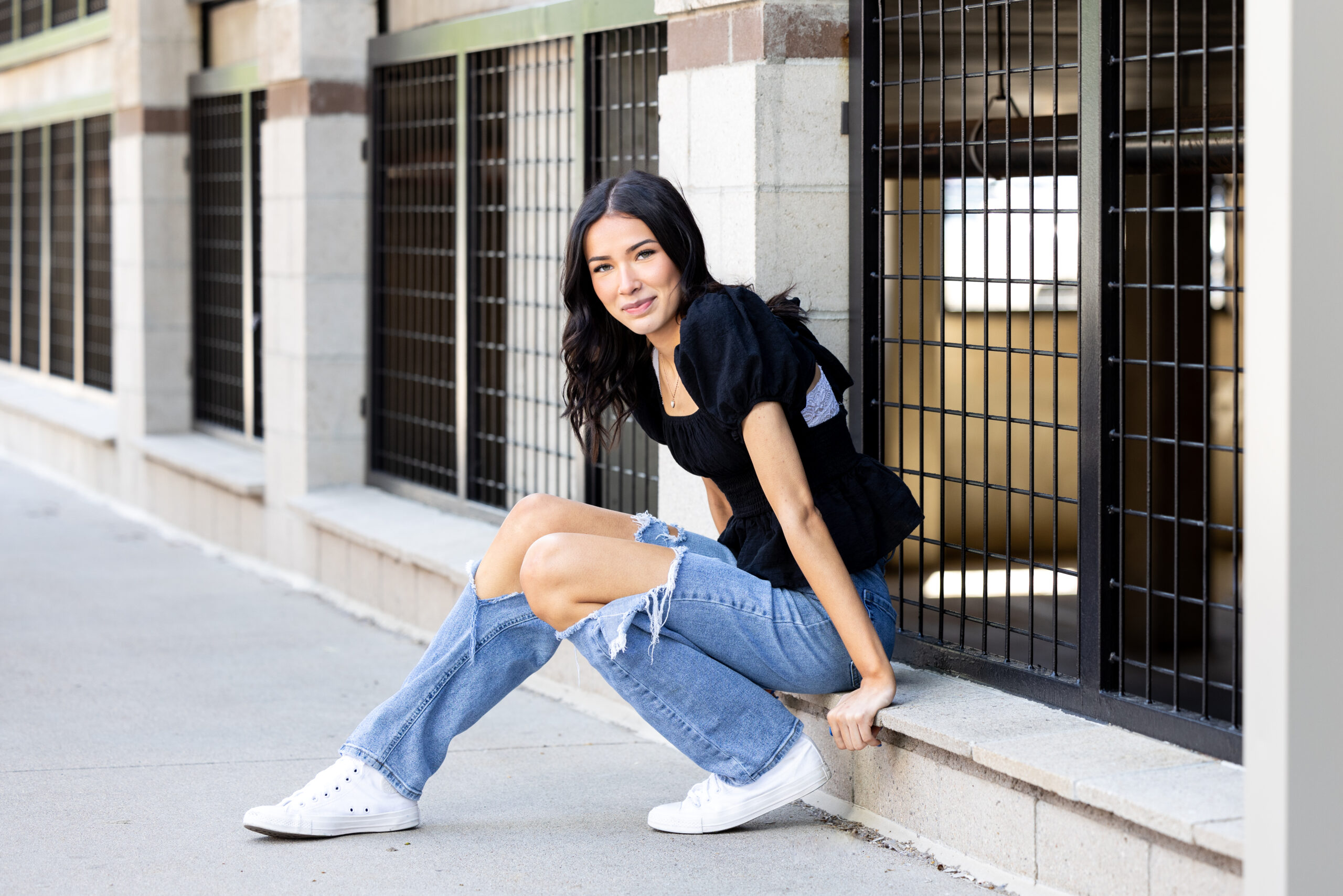 A high school senior girl is sitting on a curb outside of a parking garage in Fort Collins Colorado, leaning over and smiling for her senior pictures.