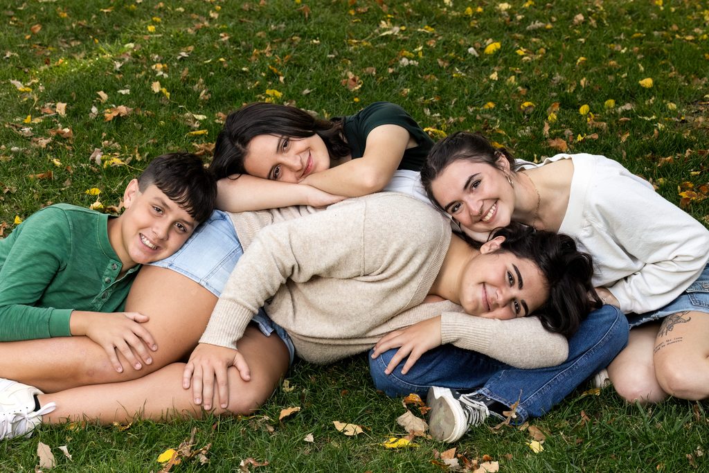 4 siblings lying on each other in yellow leaves covering green grass for their family pictures.