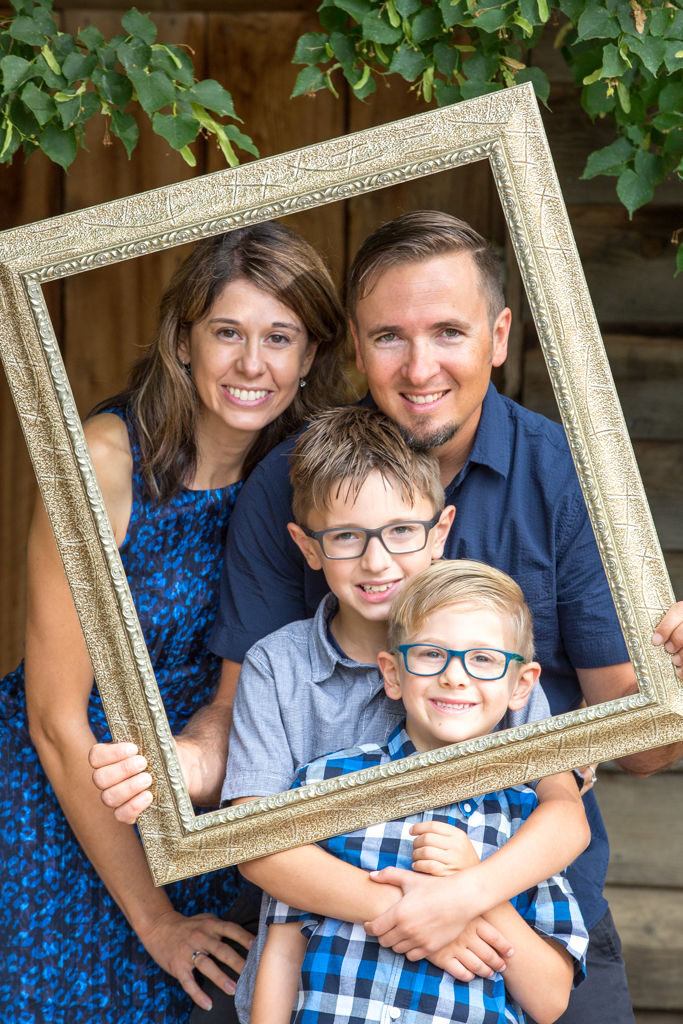 A family of 4 is standing holding a picture frame around them, smiling at the camera in Eaton Colorado with a rustic building behind them and tree branches hanging overhead.
