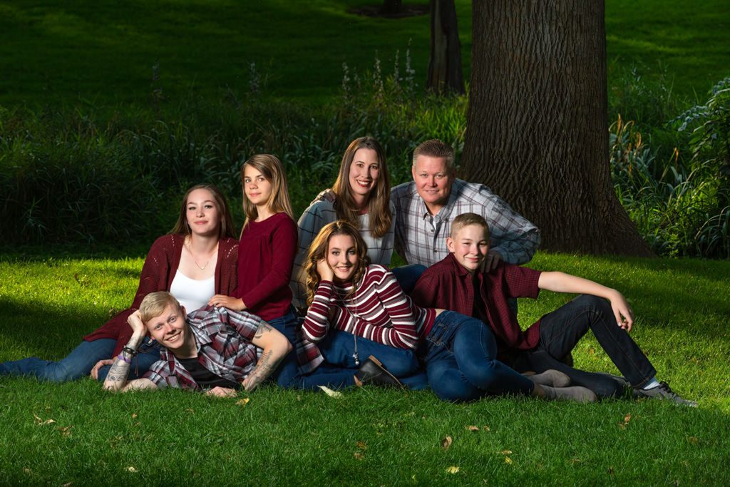 Family of 7 sitting and lying in a grassy park with a large tree behind them during their family portrait session in Greeley, Colorado.