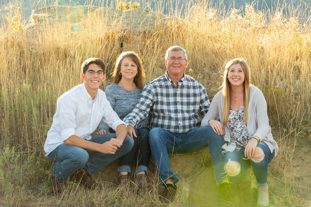 Family of 4 squatting and sitting in tall sunlit grasses smiling at the camera.