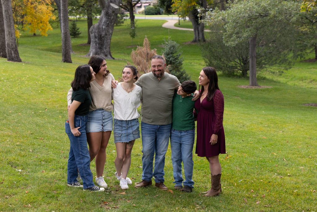 Family of 6 standing in a grassy park with trees in the background in the fall. They're smiling at each other with arms around each other.